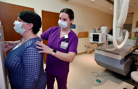 Picture of a female Nurse standing behind a female patient that is standing straight up, getting an X-RAY done.